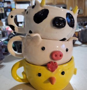 Stack of three handmade mugs with painted with the face of a duck, a pig, and a cow. 